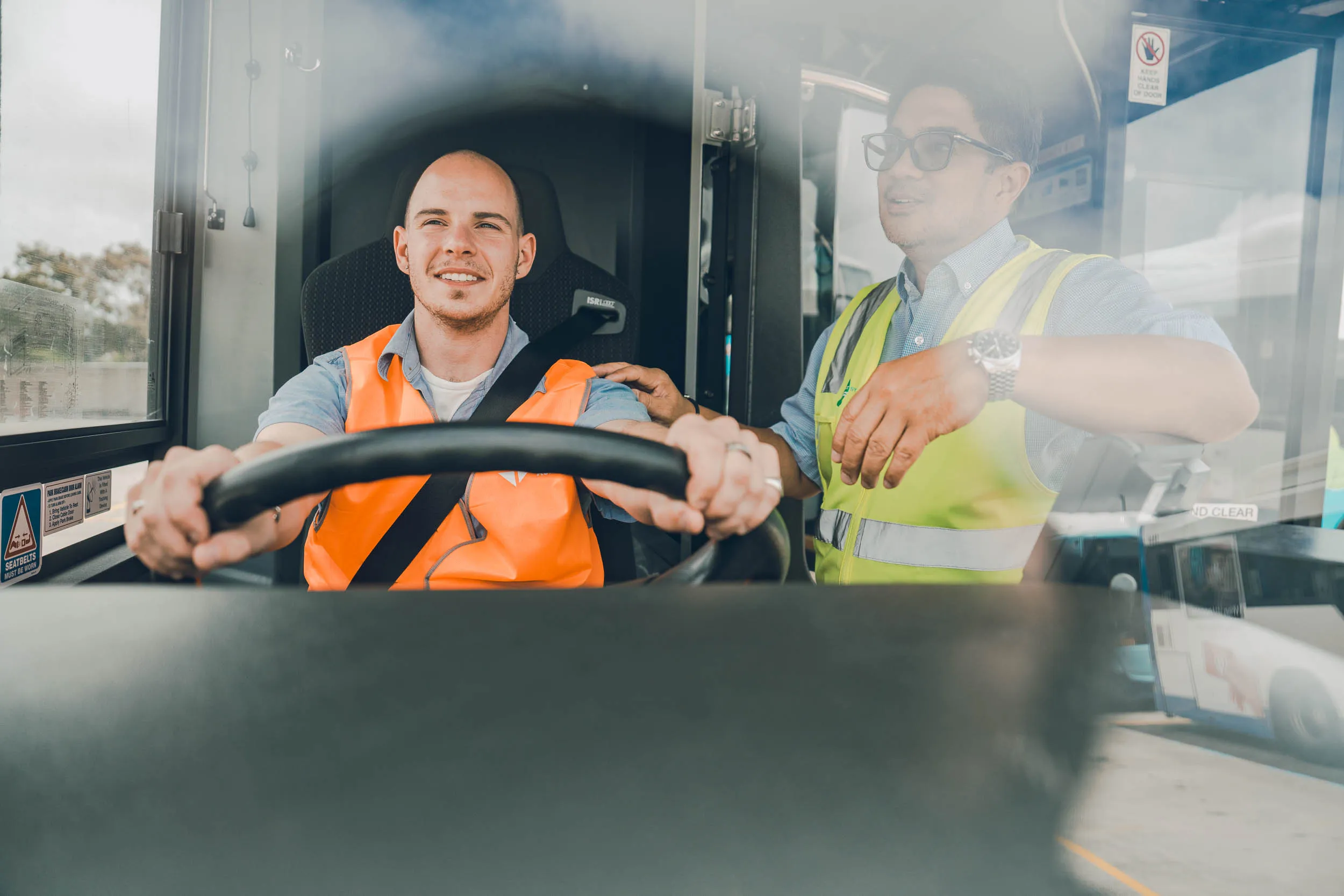 A smiling trainee bus driver sits at the wheel while an instructor in a safety vest points out directions inside the bus.