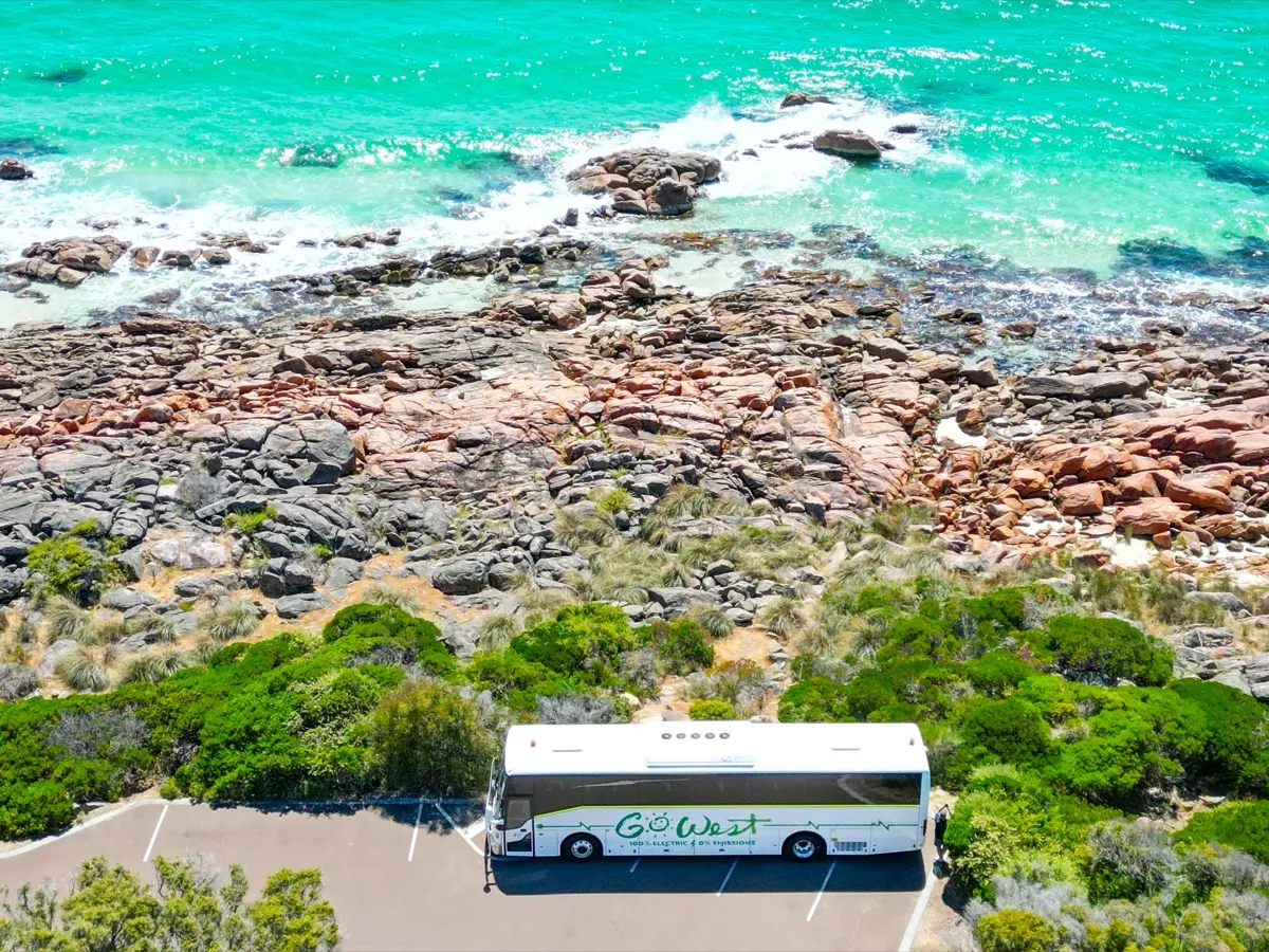 A white Go West tour bus parked in a lot beside rocky coastline and vibrant turquoise ocean water, surrounded by greenery.