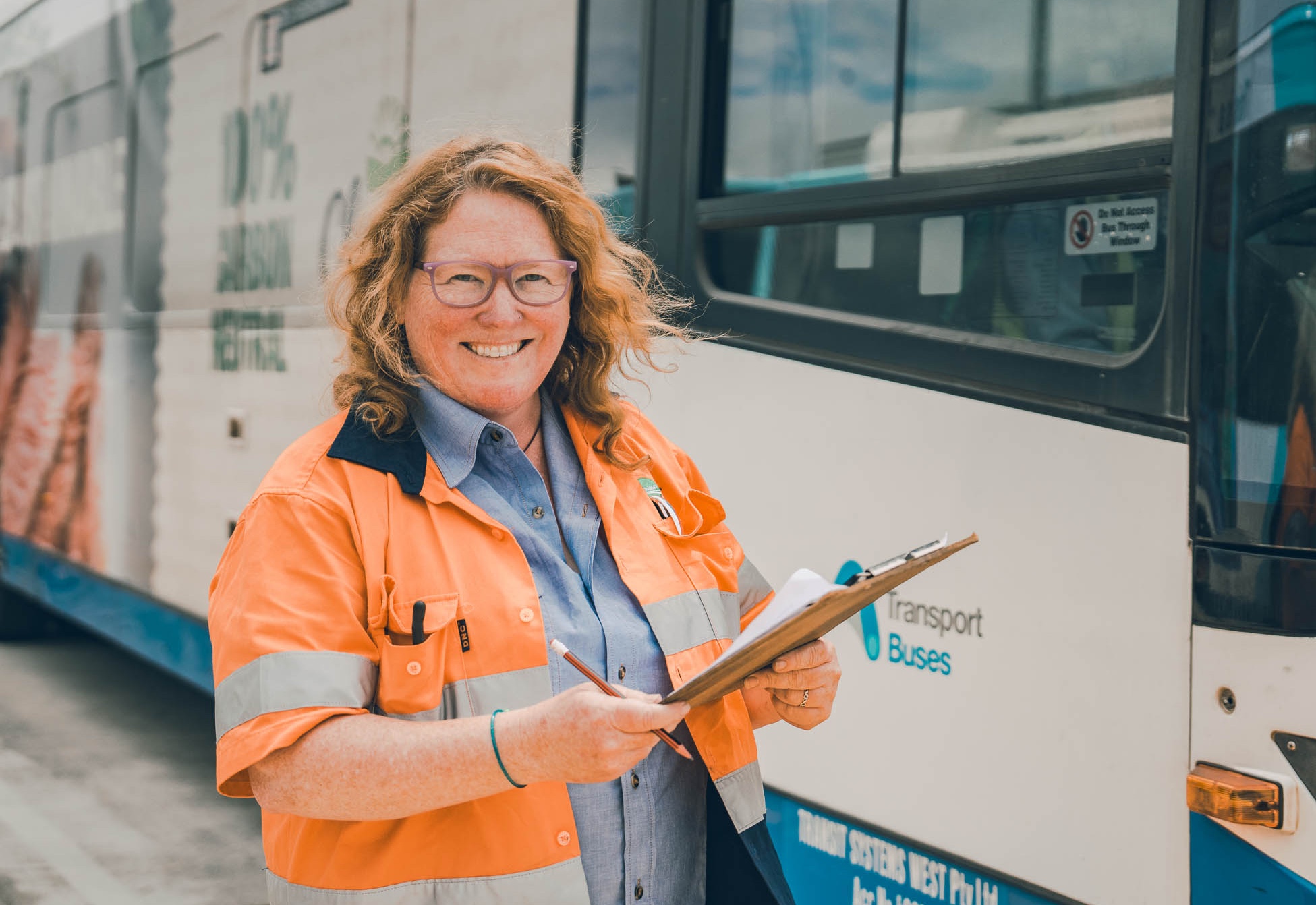 A woman wearing an orange safety jacket and glasses stands smiling with a clipboard in hand beside a Transport Buses vehicle.