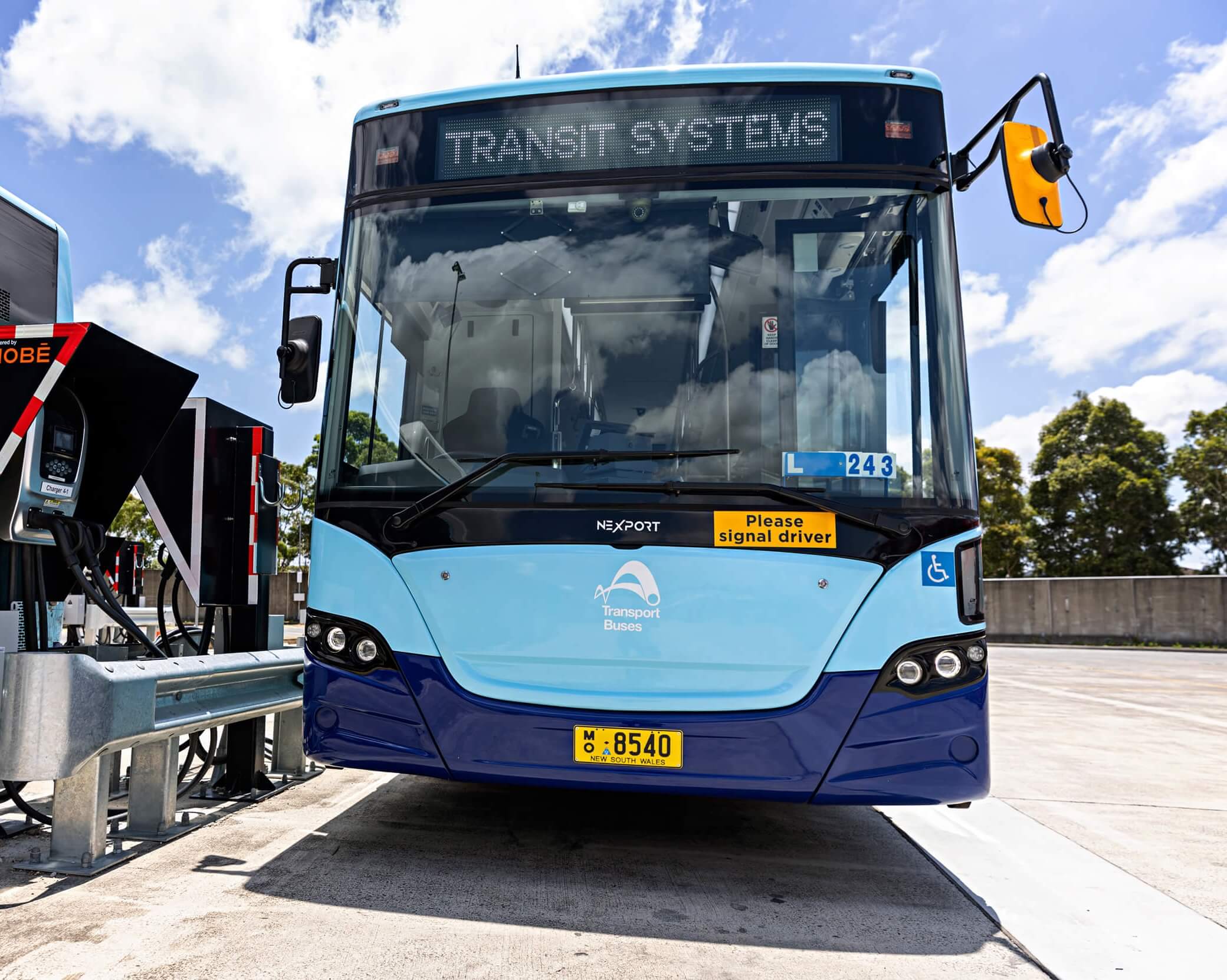 A light blue Transit Systems bus parked at a charging station under a sunny sky, with electric charging equipment visible on the left.