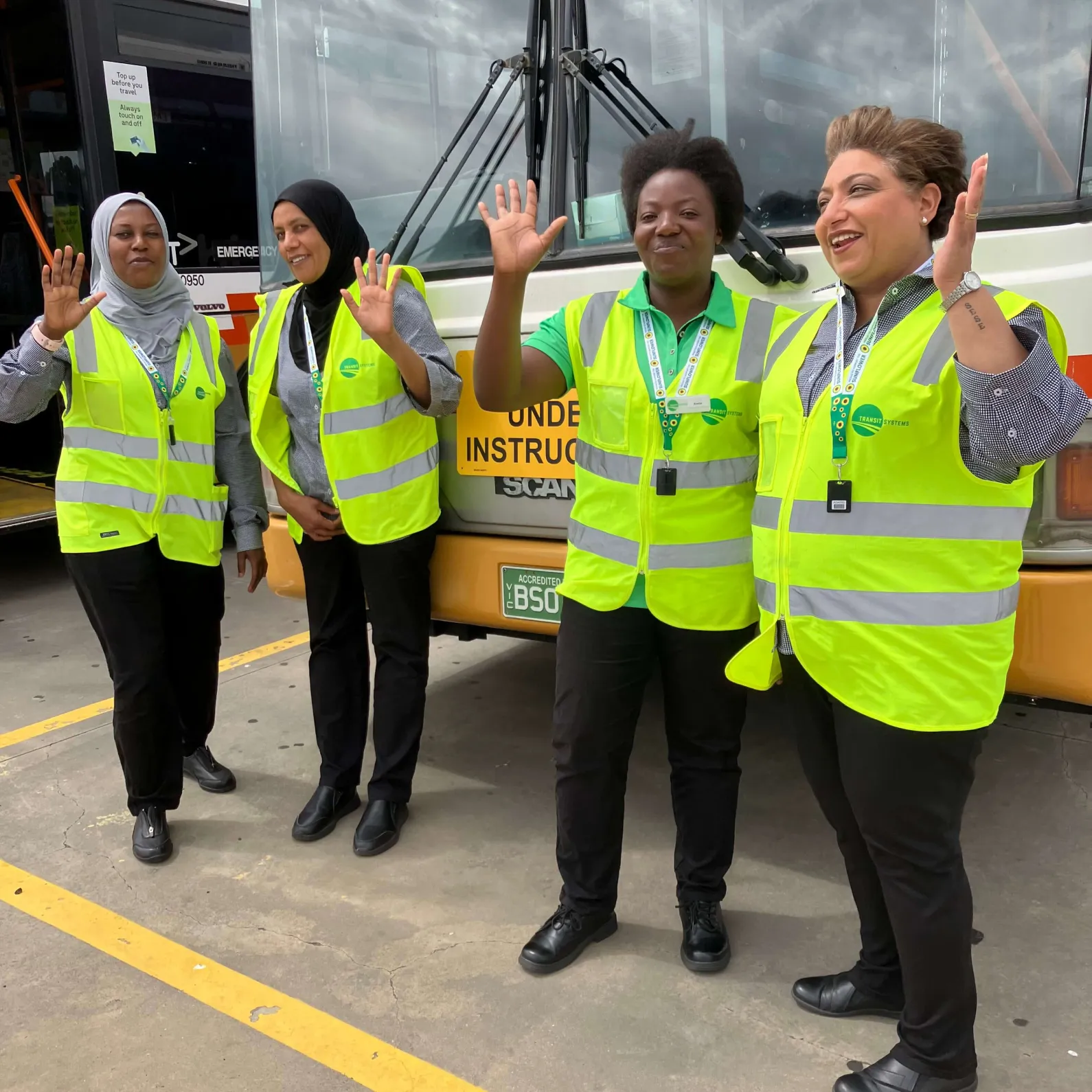 our women wearing high-visibility vests smiling and waving while standing in front of a bus in a parking lot.