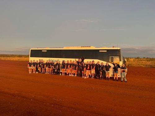 A large group of people standing in front of a white coach bus on a red dirt road, with golden sunlight and open outback landscape in the background.