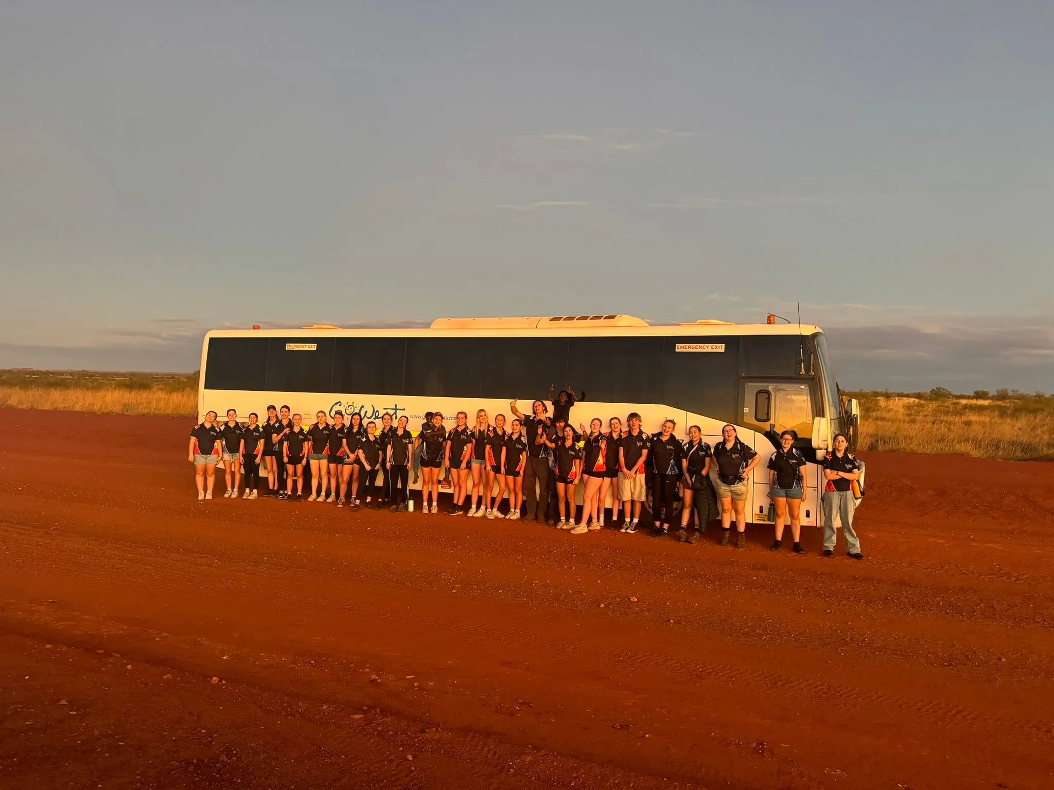A large group of people standing in front of a white coach bus on a red dirt road, with golden sunlight and open outback landscape in the background.