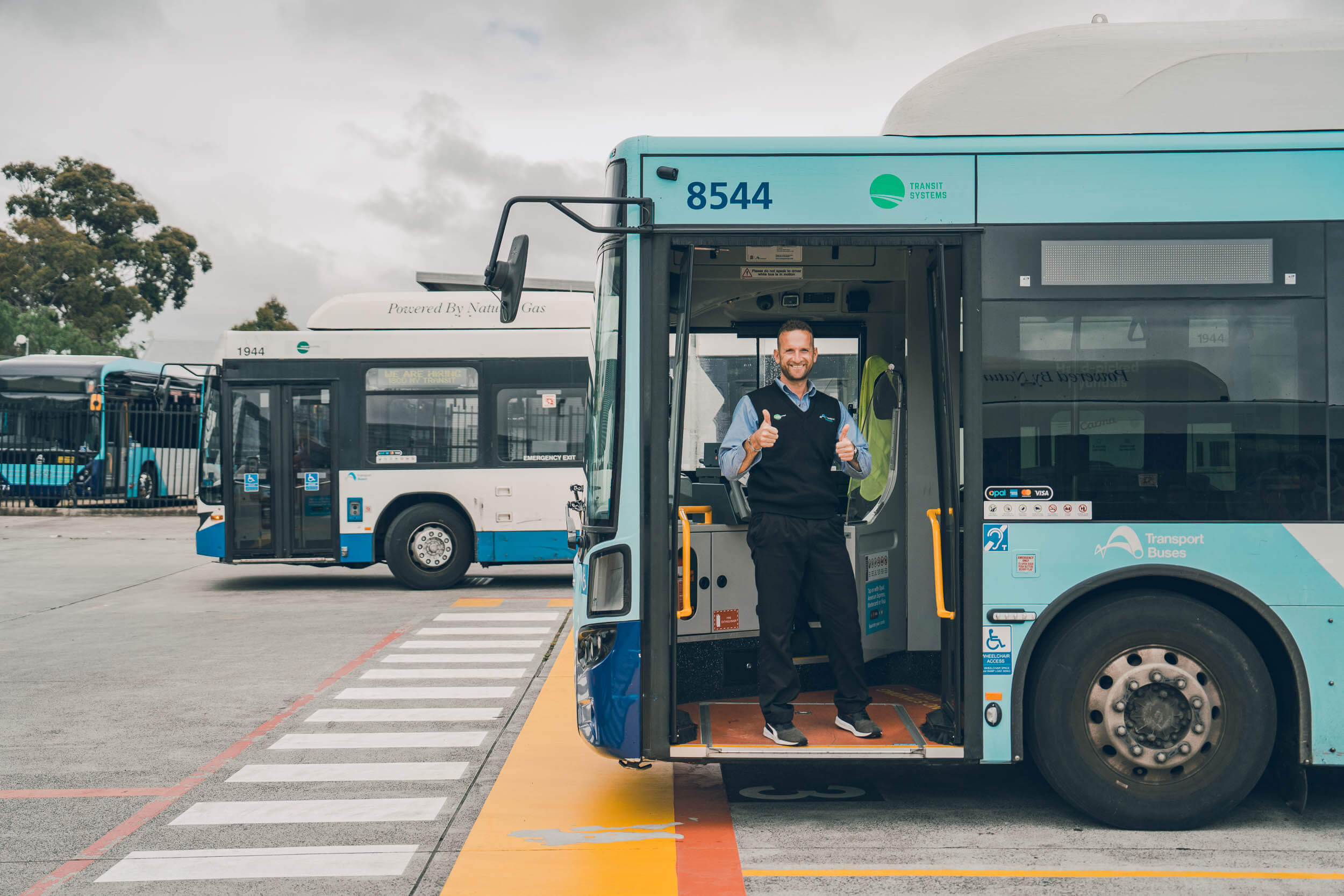A smiling bus driver giving two thumbs up while standing in the doorway of a Transit Systems bus at a bus depot, with other buses and trees in the background.