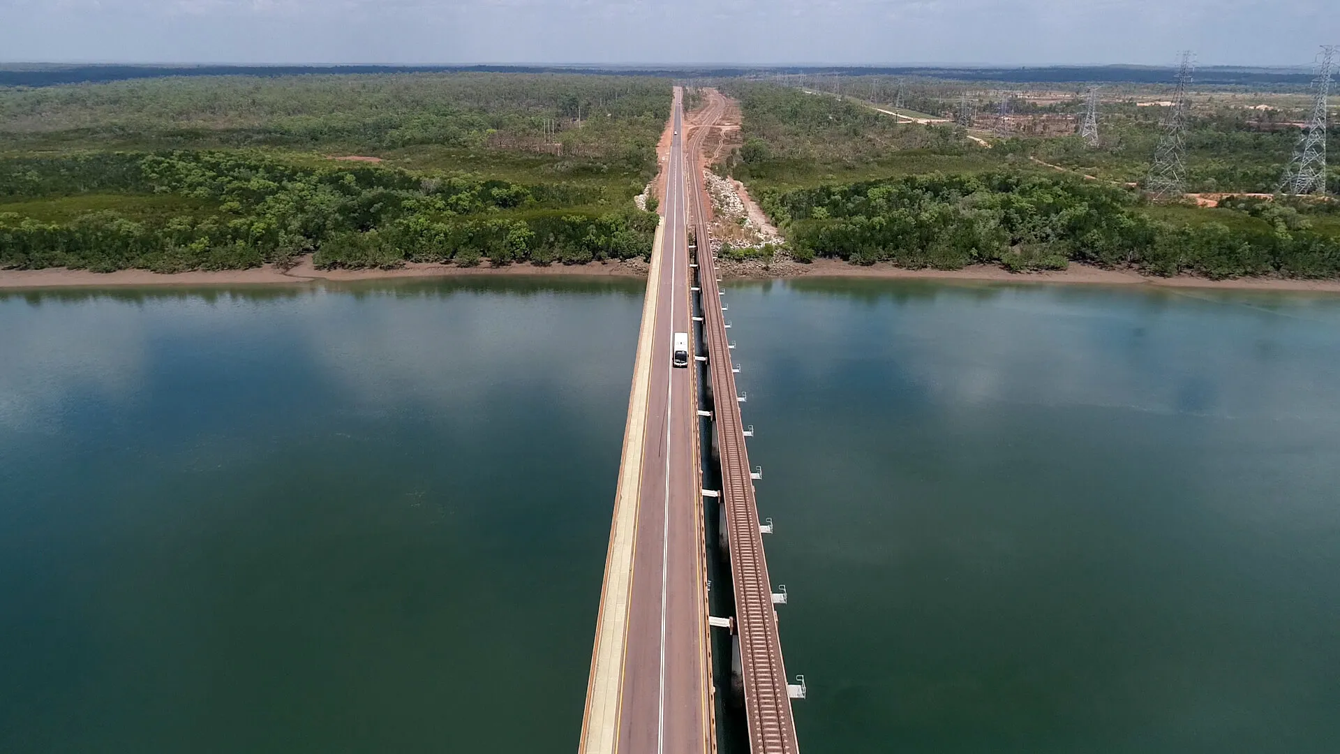 A long, straight bridge crossing over a wide river with a single vehicle traveling across, surrounded by dense green forest and power lines in the distance.