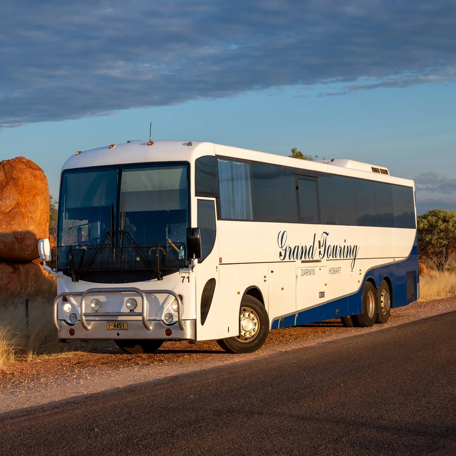 White and blue Grand Touring coach bus parked on a rural road near large red rocks under a partly cloudy sky.