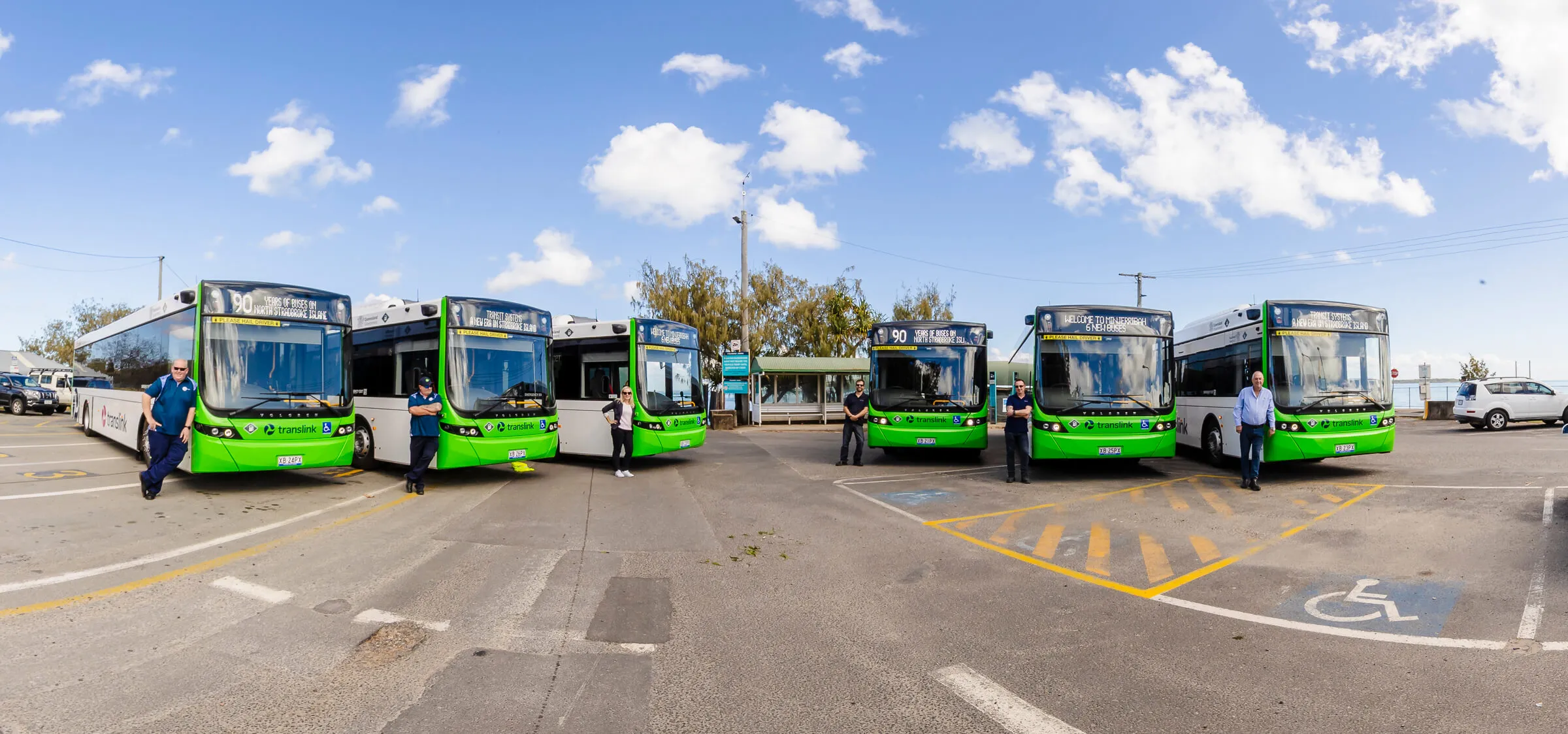 A row of bright green buses is parked in a lot under a blue sky with scattered clouds, with several bus drivers standing beside or in front of the vehicles.