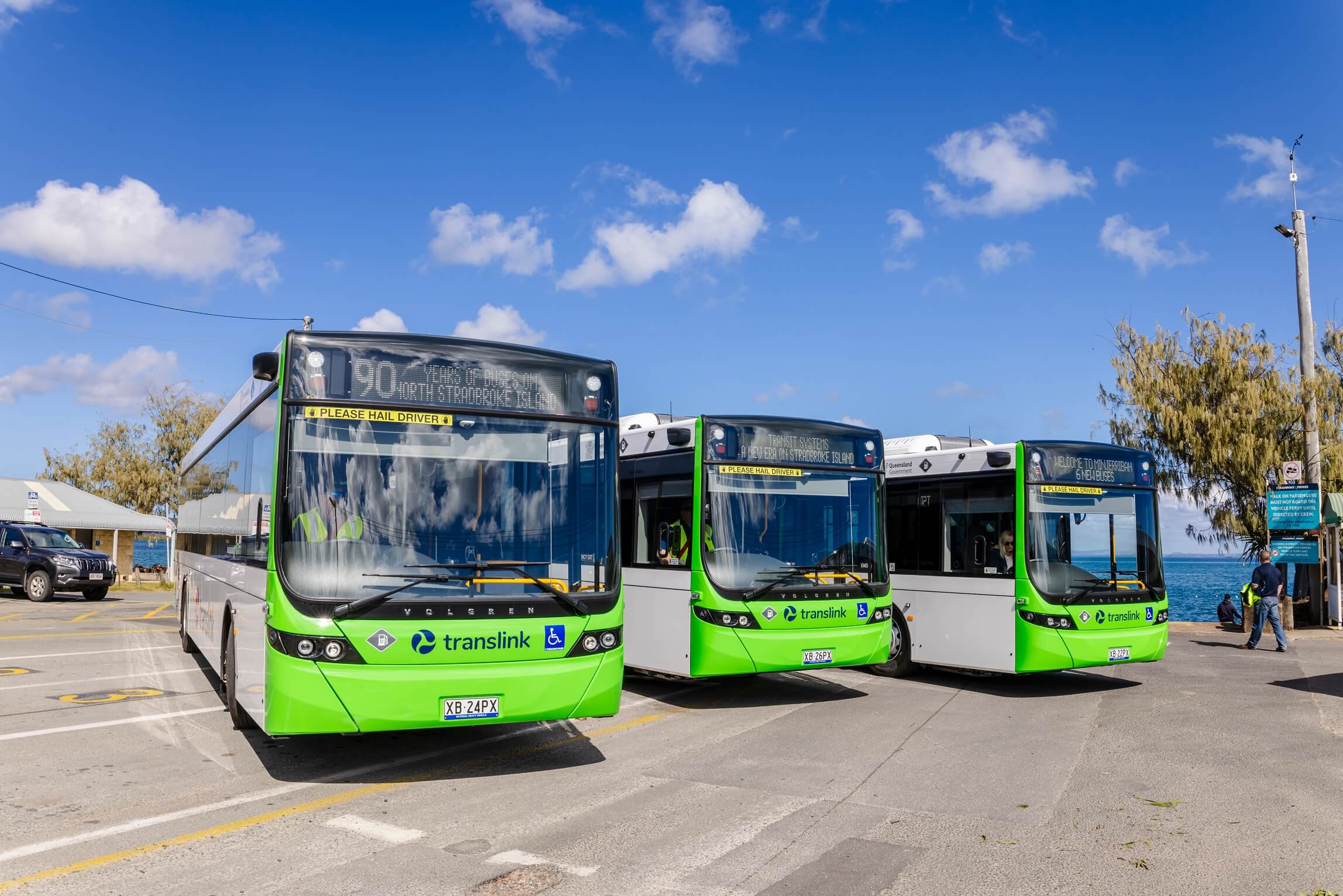 Three green and white TransLink buses parked near the coast under a blue sky.