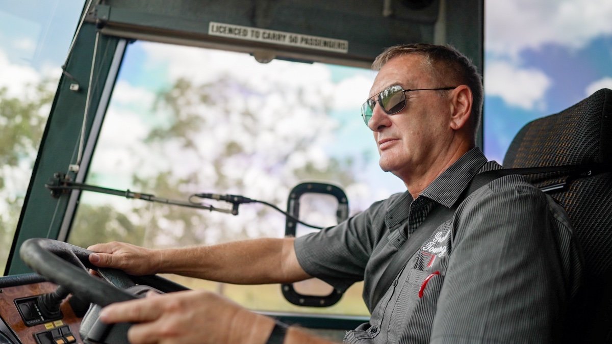 A bus driver wearing sunglasses and a grey shirt is focused on the road while driving, with trees and blue sky visible through the windshield.