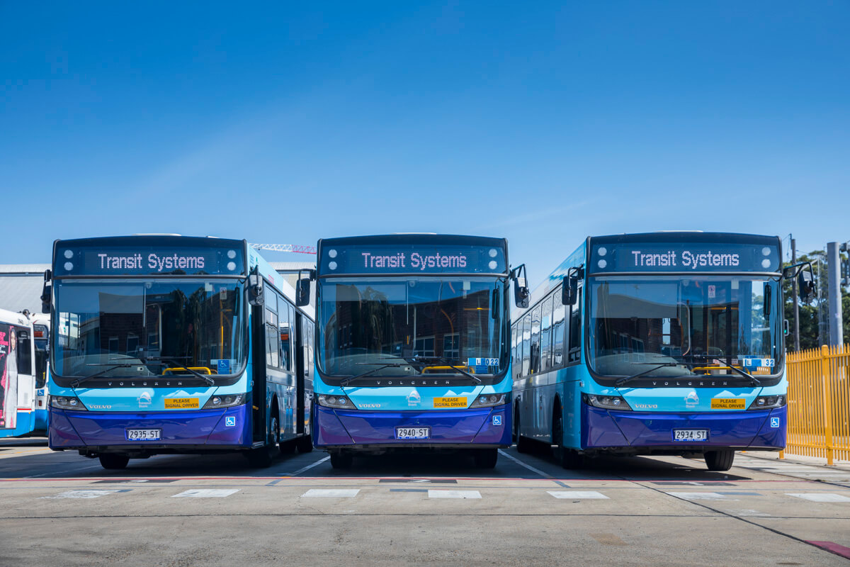 Three blue and purple city buses labeled "Transit Systems" are parked side by side in a bus depot under a clear blue sky.