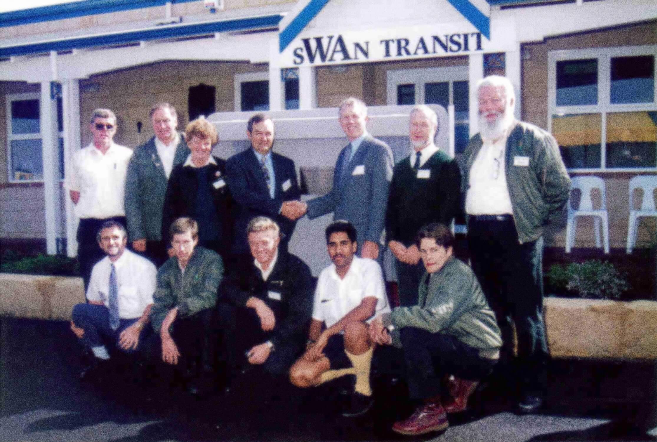 A group of people pose for a photo outside a building with a sign that reads "Swan Transit," with two individuals in the center shaking hands.