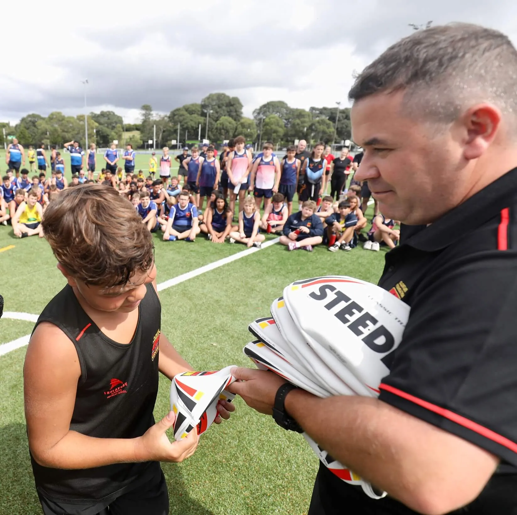 A young boy receives a rugby ball from an adult coach during a sports event, with a group of children sitting and standing in the background on a grassy field.