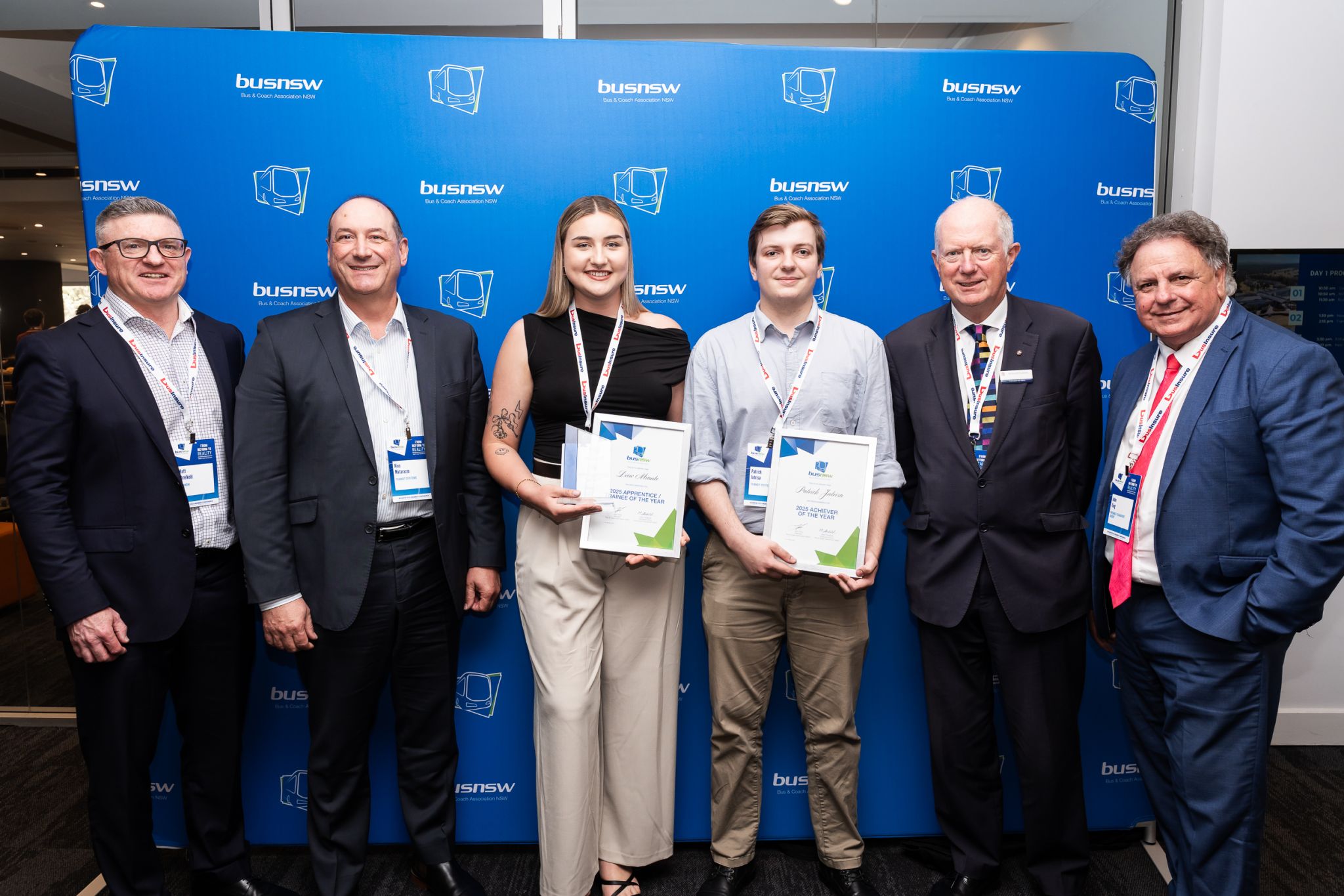 Group photo at a BusNSW event with award winners holding certificates, standing in front of a branded blue backdrop with six professionally dressed individuals.