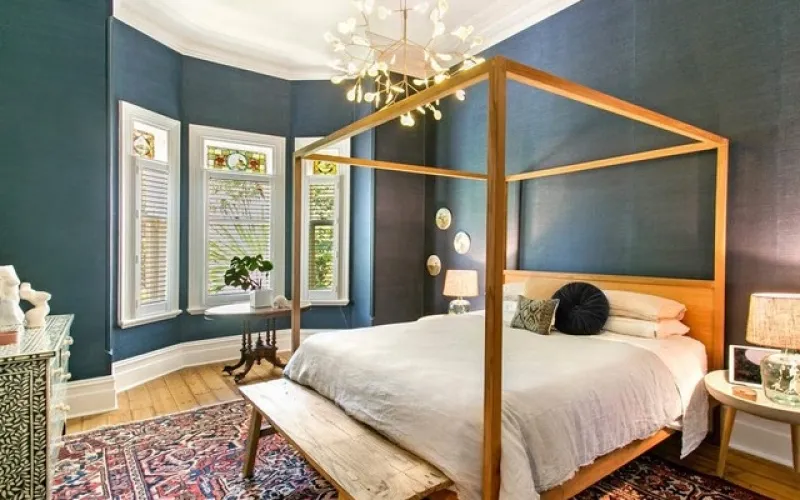 Bedroom with wooden four-poster bed, white bedding, blue walls, bay windows, and colorful patterned rug.