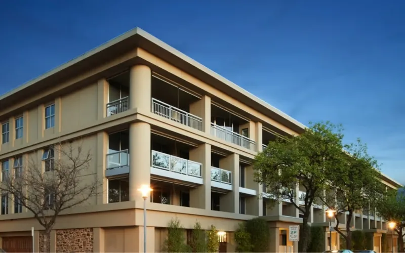 Modern beige apartment building with balconies and trees under a clear blue sky at dusk.