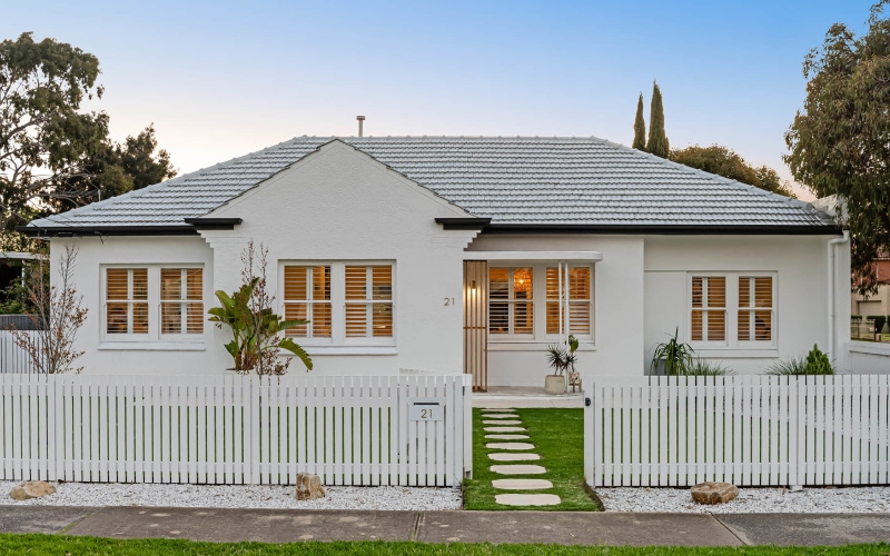 White single-story house with grey tiled roof, white picket fence, and a grass path leading to the entrance.