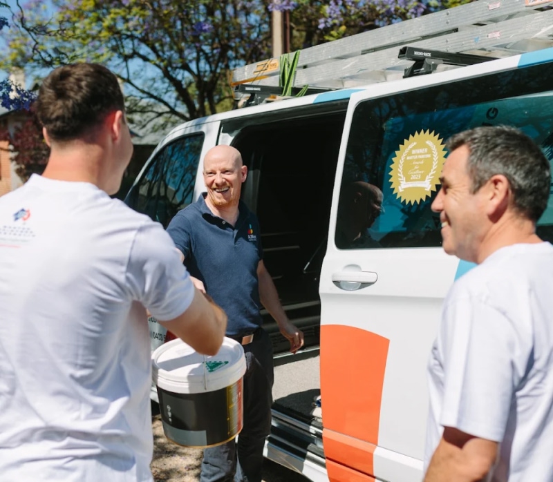 Three men smiling near a work van; one man hands a paint bucket to another outdoors.