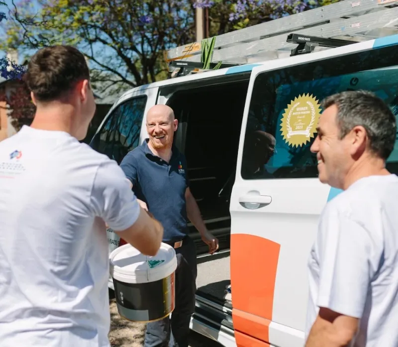 Three men smiling near a work van; one man hands a paint bucket to another outdoors.