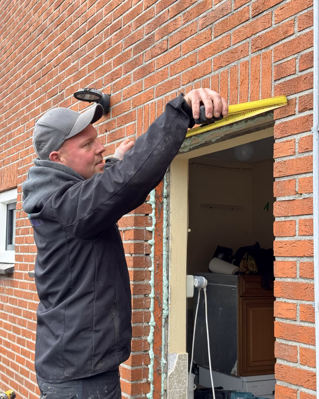 Man in jas en pet meet met gele meetlat boven deuropening in rode bakstenen muur.
