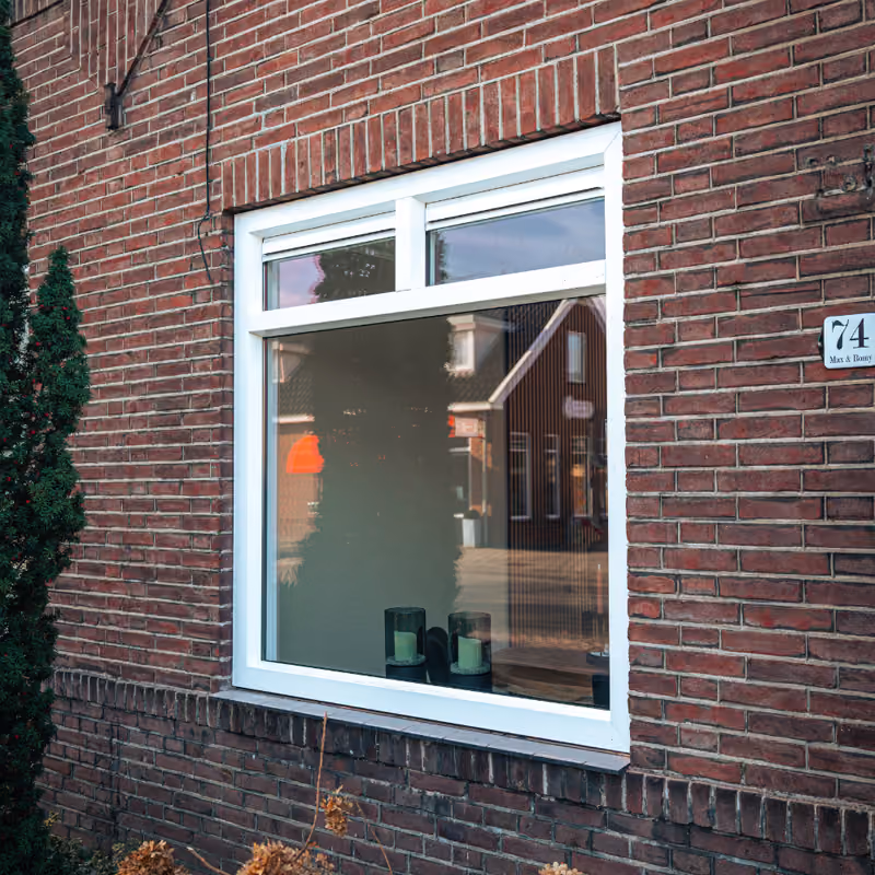 Wit kozijn met groot glas in een roodstenen gevel met reflectie van huizen in het raam.