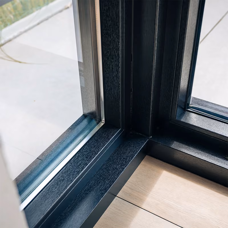 Detail van een antracietkleurig kozijn met vast glas en hoekverbinding op houten vloer.