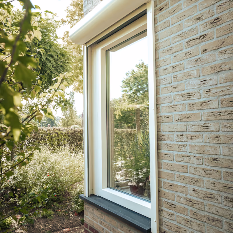 Wit raampje met glas in een bakstenen muur, omringd door groen en planten buiten.