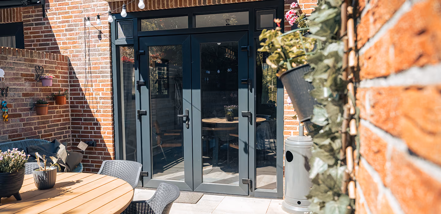 Open antraciet schuifdeuren leiden naar een terras met ronde houten tafel, stoelen en planten.
