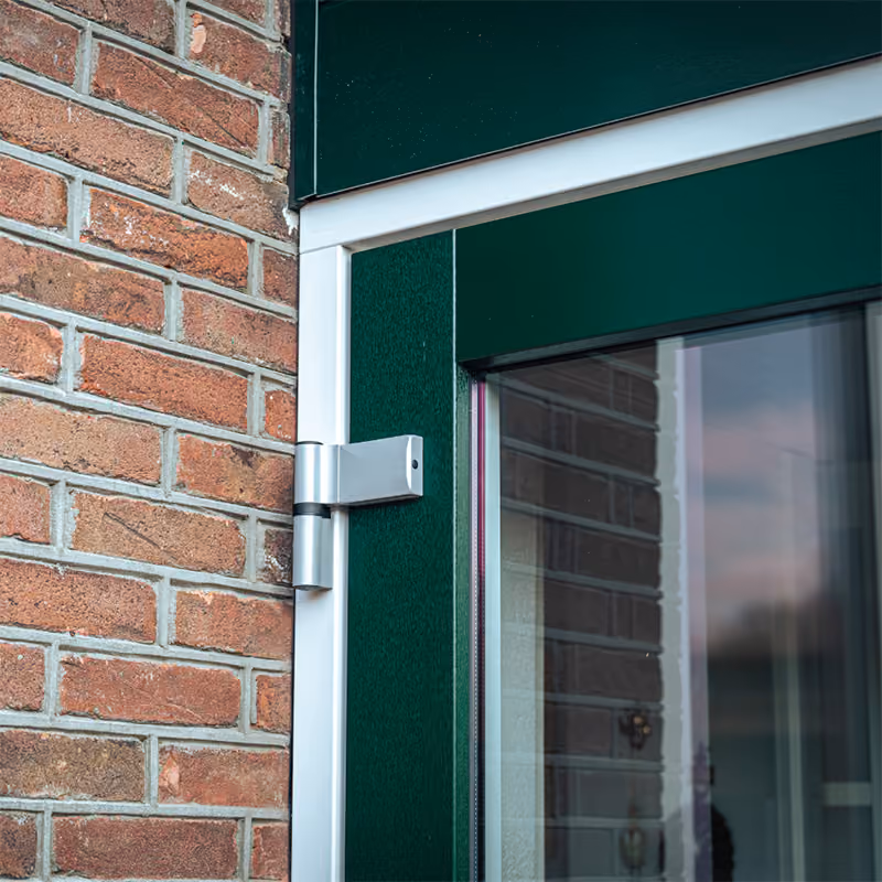Detailfoto van een openende groene deur met zilver scharnier tegen een bakstenen muur.