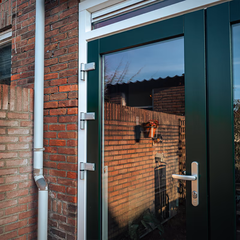 Donkergroene buitendeur met glas en metalen scharnieren tegen een gevel van rode bakstenen.