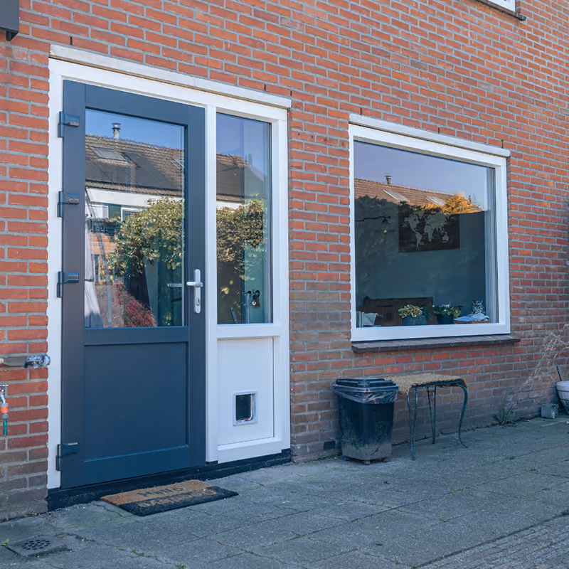 Achterdeur met kattenluik in antraciet en wit naast een raam in een bakstenen gevel.