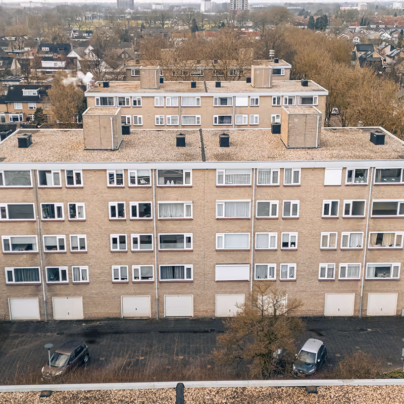 Luchtfoto van een groot appartementsgebouw met parkeerplaatsen, bomen en woonwijk op de achtergrond.