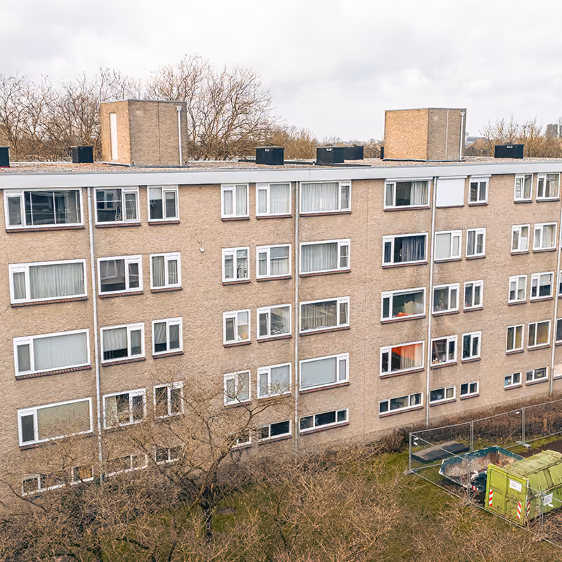 Flatgebouw met meerdere kunststof kozijnen en een groene afvalcontainer in de voorgrond.