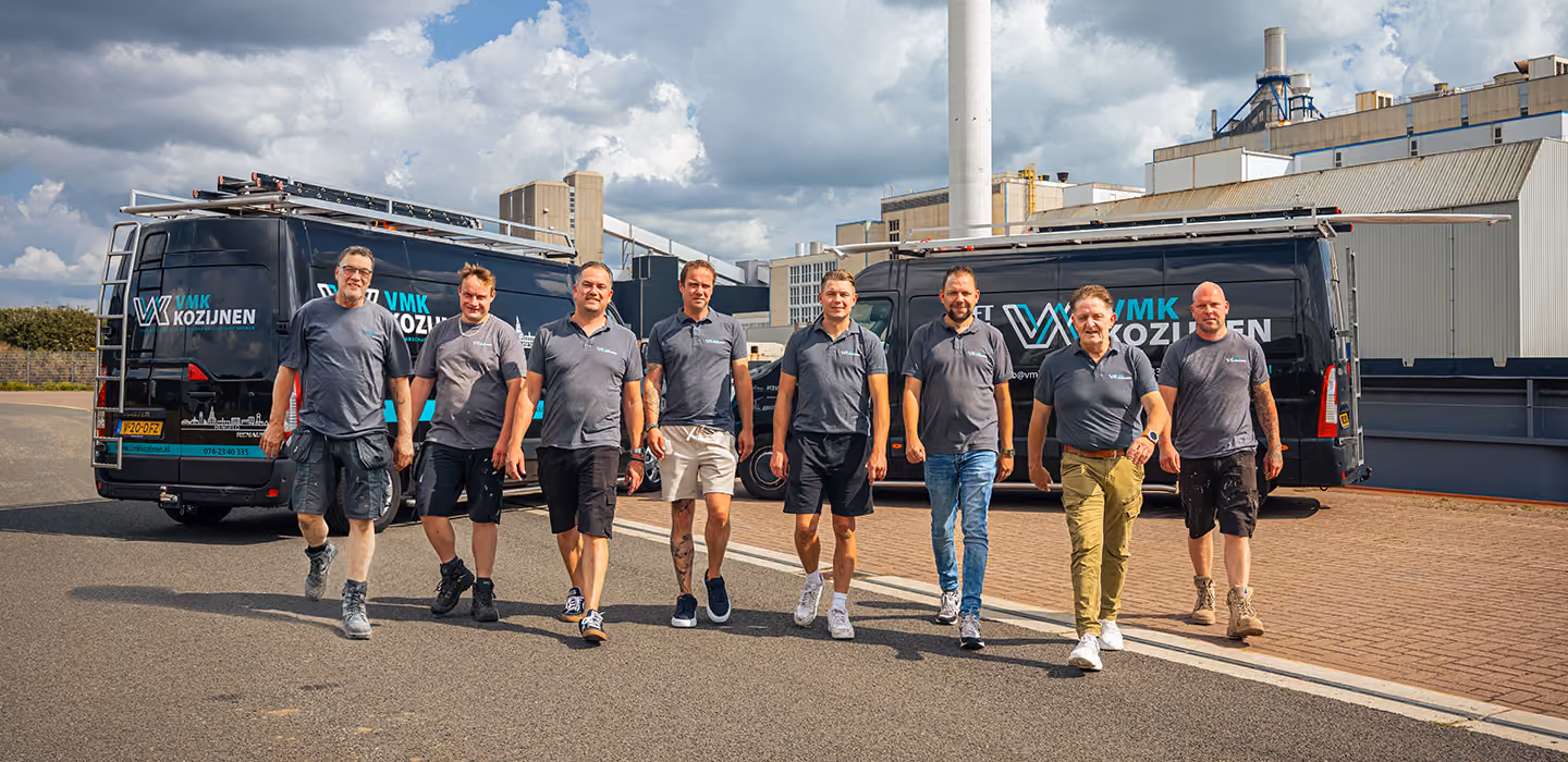 Team van acht mannen in grijze shirts loopt voor twee zwarte bedrijfsbussen met VMK Kozijnen-logo.