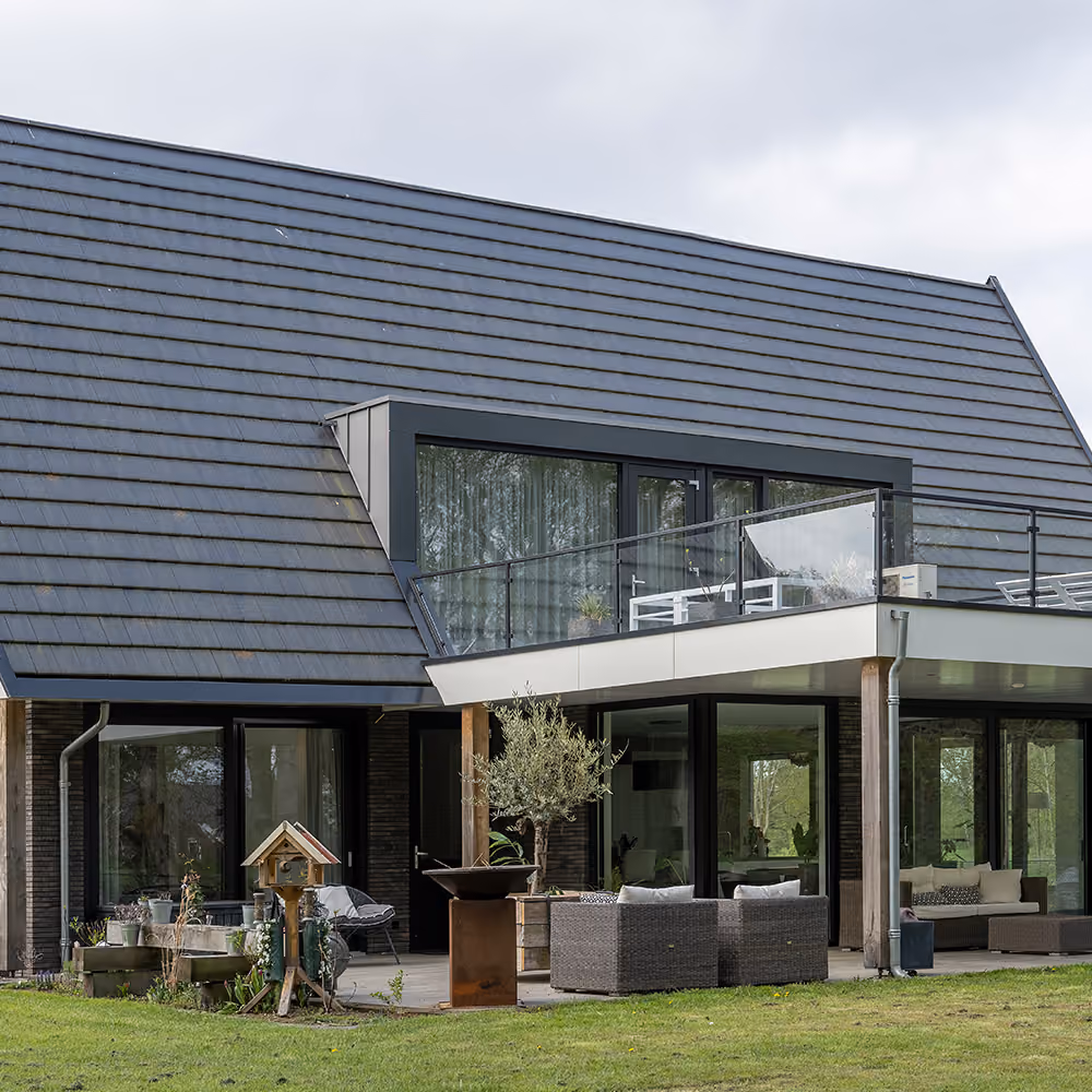 Moderne villa met zwart dak en grote ramen, een balkon met glazen balustrade en een terras met tuinmeubilair op een groene grasmat.