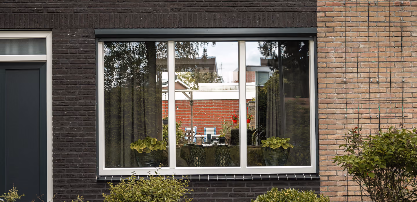 Grote witte schuiframen in een huisgevel van donkere en lichte baksteen met groene planten buiten en een weerspiegeling van een tuin en omringende huizen in het glas.