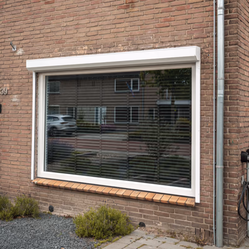 Groot rechthoekig raam met witte omlijsting in een bakstenen muur, met zwarte jaloezieën en de weerspiegeling van een straat met auto's en huizen erop.