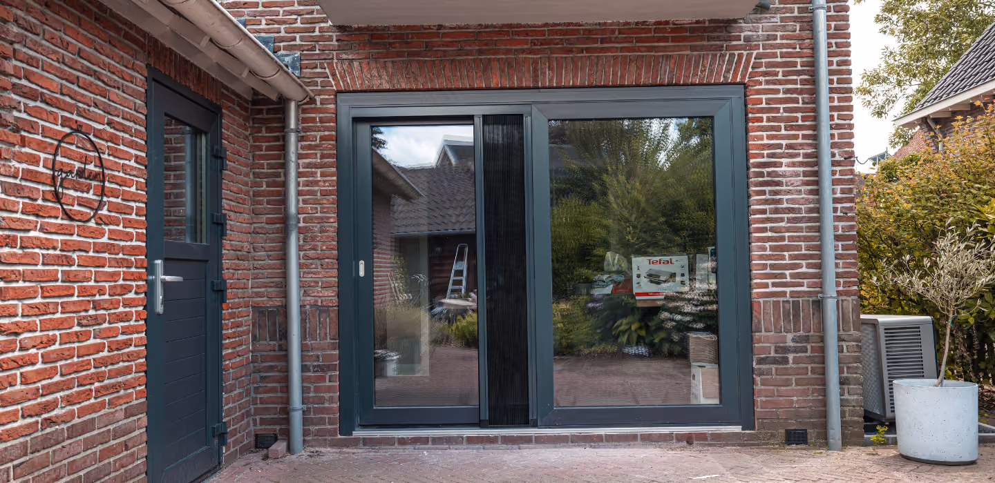 Twee-delige schuifpui met donkere kozijnen ingebouwd in een gevel van rode bakstenen, met een donkere deur links en plantenbak rechts.