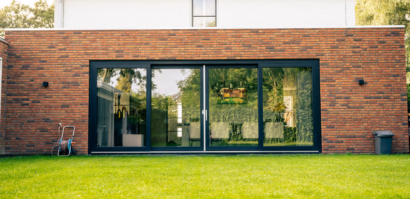 Moderne tuin met strak groene grasmat en een lage woning met een brede, vierdelige glazen schuifpui in donkere omlijsting tegen een gevel van baksteen.