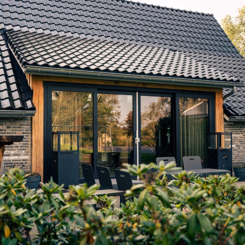 Moderne huisgevel met een zwarte geglazuurde dakpan, grote glazen schuifdeuren ingesloten door een houten frame, en buitenterras met zwarte stoelen en planten op de voorgrond.