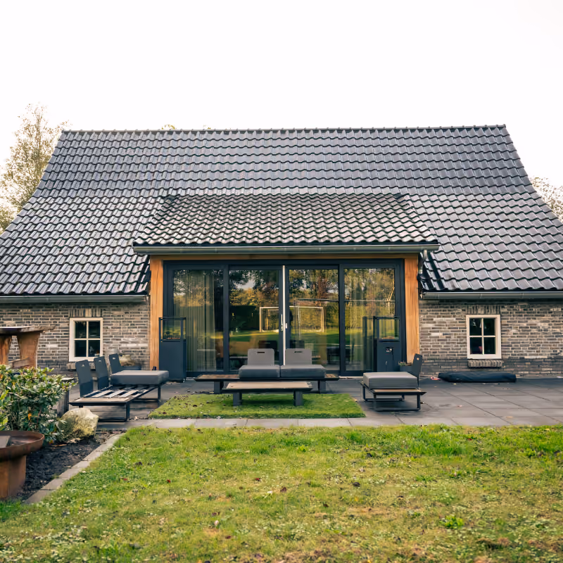Moderne achterzijde van een huis met een grote glazen schuifpui, gepleisterde bakstenen muren en een zwart dak met golvende dakpannen, met een terras waarop stijlvolle loungebank en stoelen staan en een groen gazon op de voorgrond.