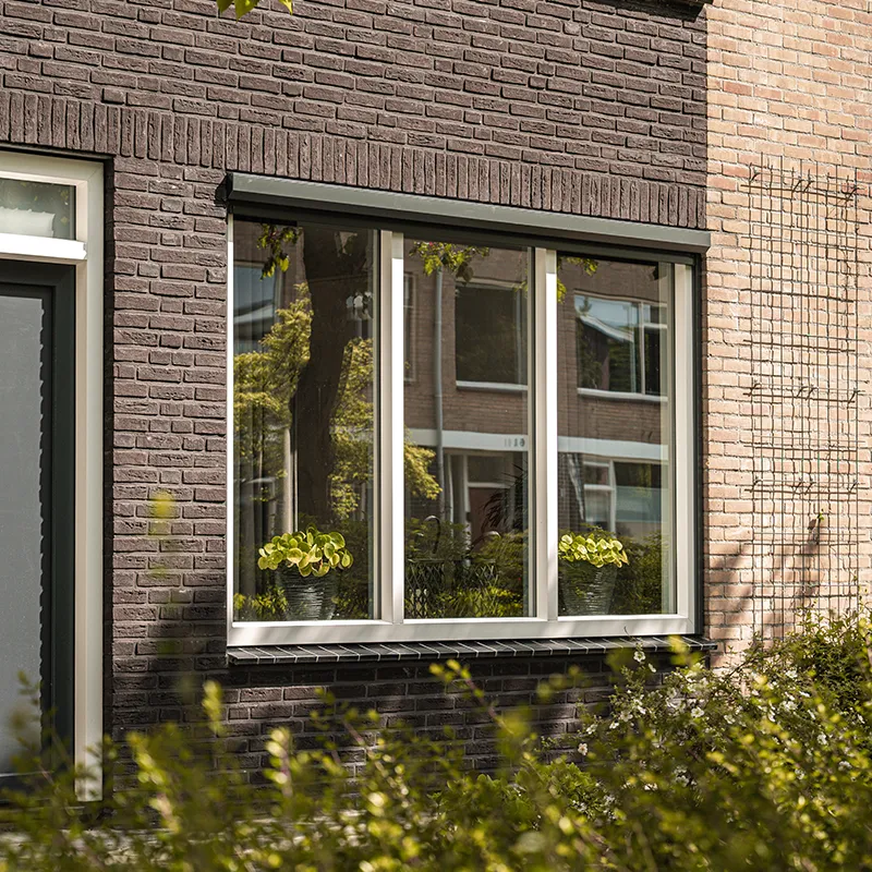 Kunststof raam met drie vakken en een grijswit kozijn in een donkere bakstenen gevel, naast een voordeur met glasstrook