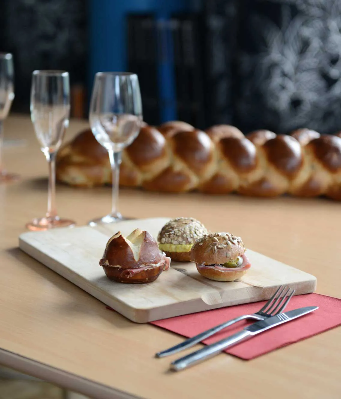 Drei belegte Mini-Brötchen auf einem Holzbrett, daneben leere Sektgläser und Besteck auf einem rot gefalteten Serviettentuch; im Hintergrund ein großer, geflochtener Hefezopf.