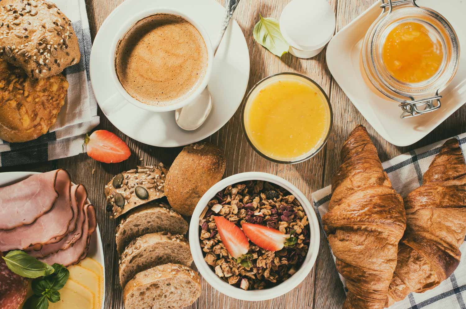 Holztisch mit diversen Brunch Leckereien wie Café, Orangensaft, Gipfeli, Brot, Müesli, Schinken, Früchte.