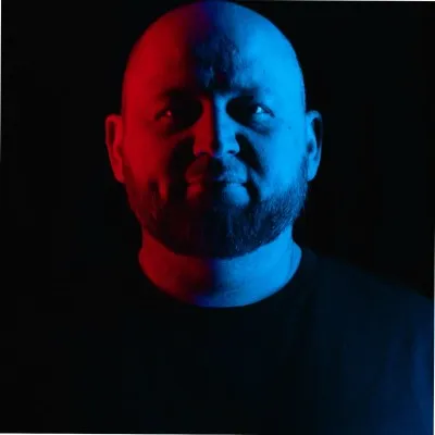 Man with bald head and beard lit dramatically in red and blue light against a black background.