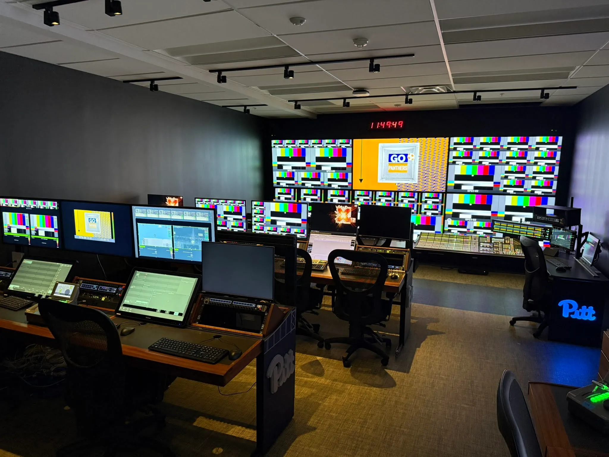 Wide shot of a modern multi-station broadcast control room with large monitor wall displaying test patterns and Pitt branding.