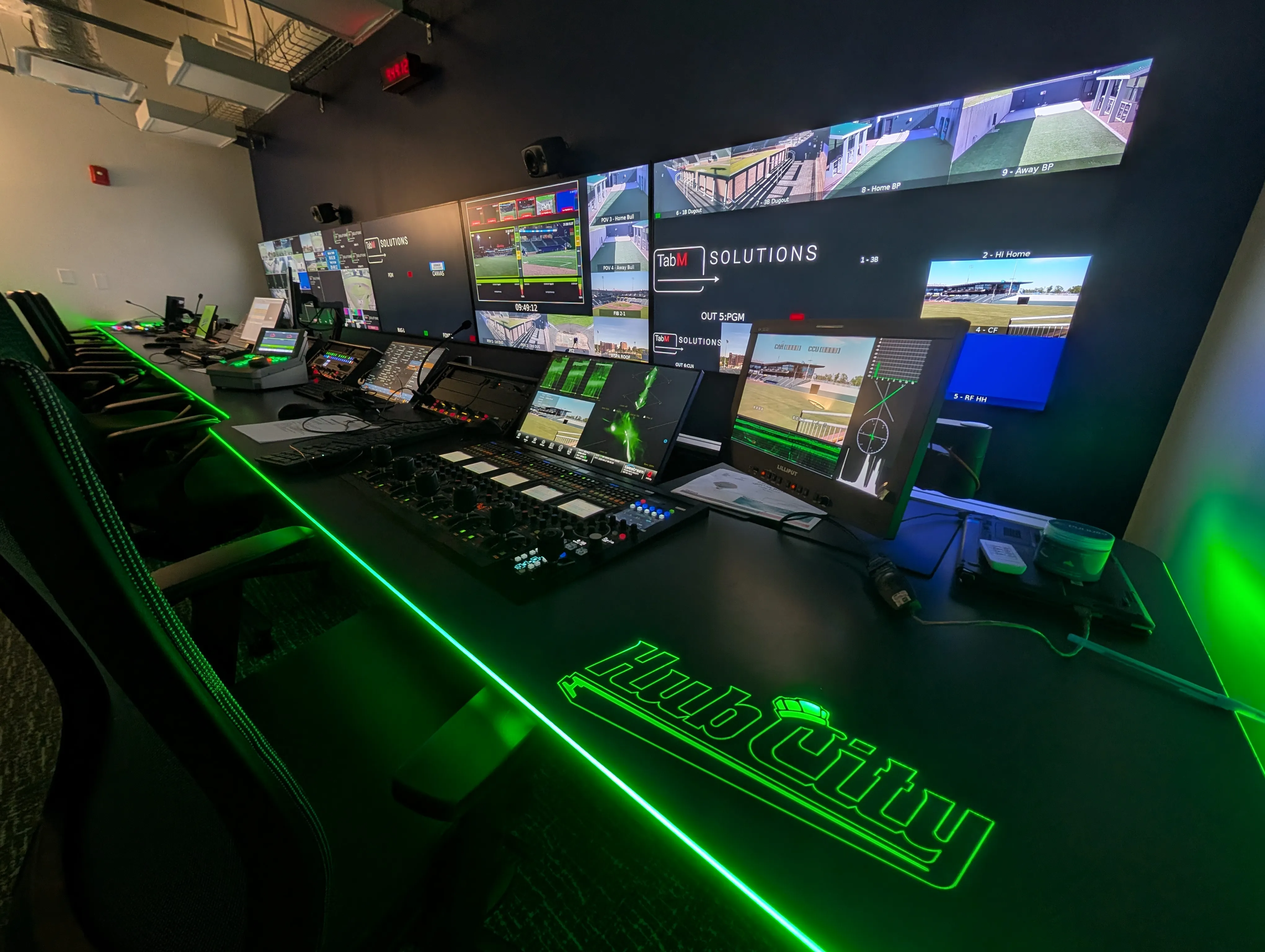 Modern broadcast control room with multiple monitors displaying baseball field views and video controls, illuminated by green LED lighting on the desk labeled Hub City.