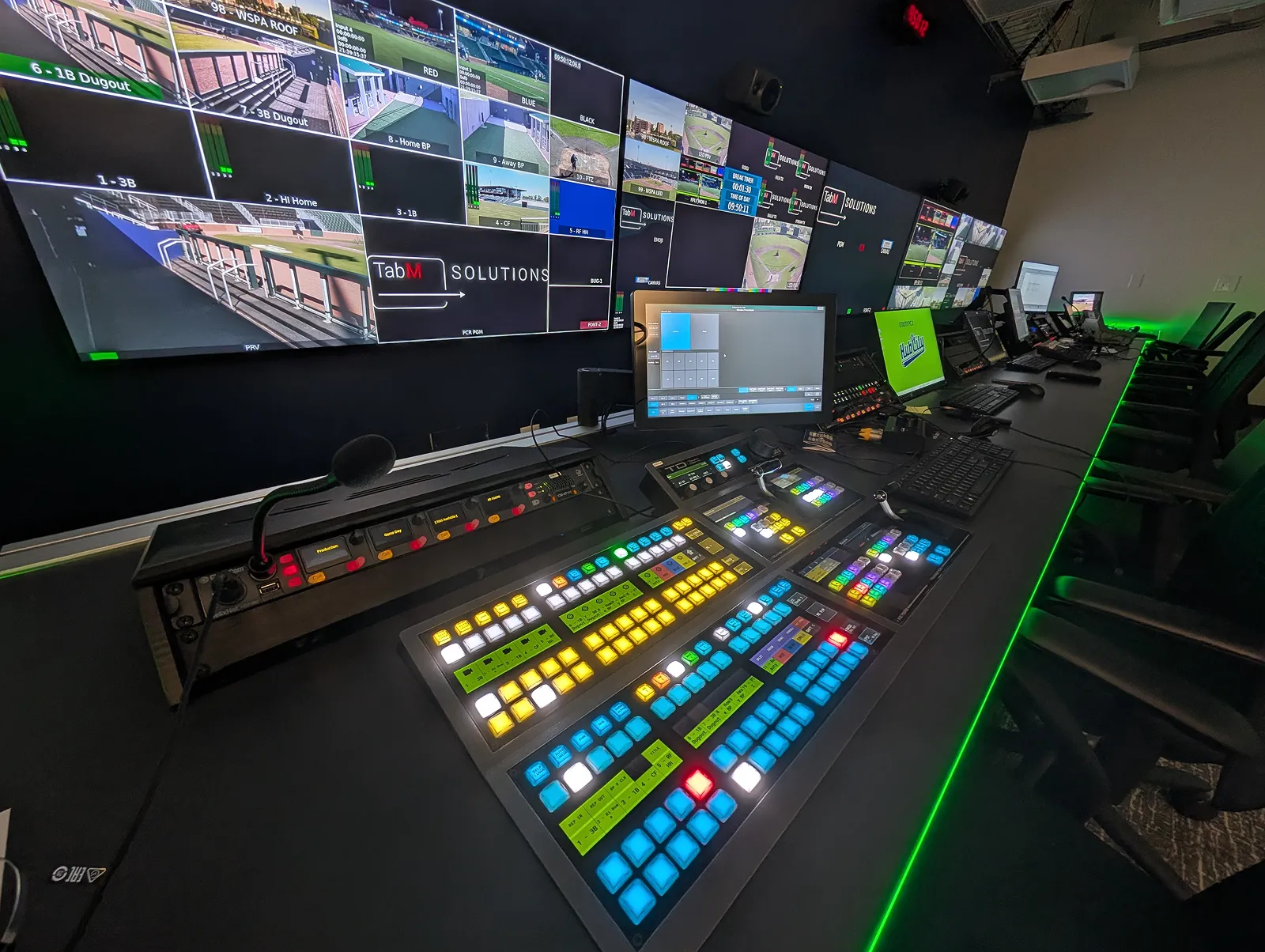 Broadcast control room with illuminated video switcher and multiple screens displaying sports field footage.