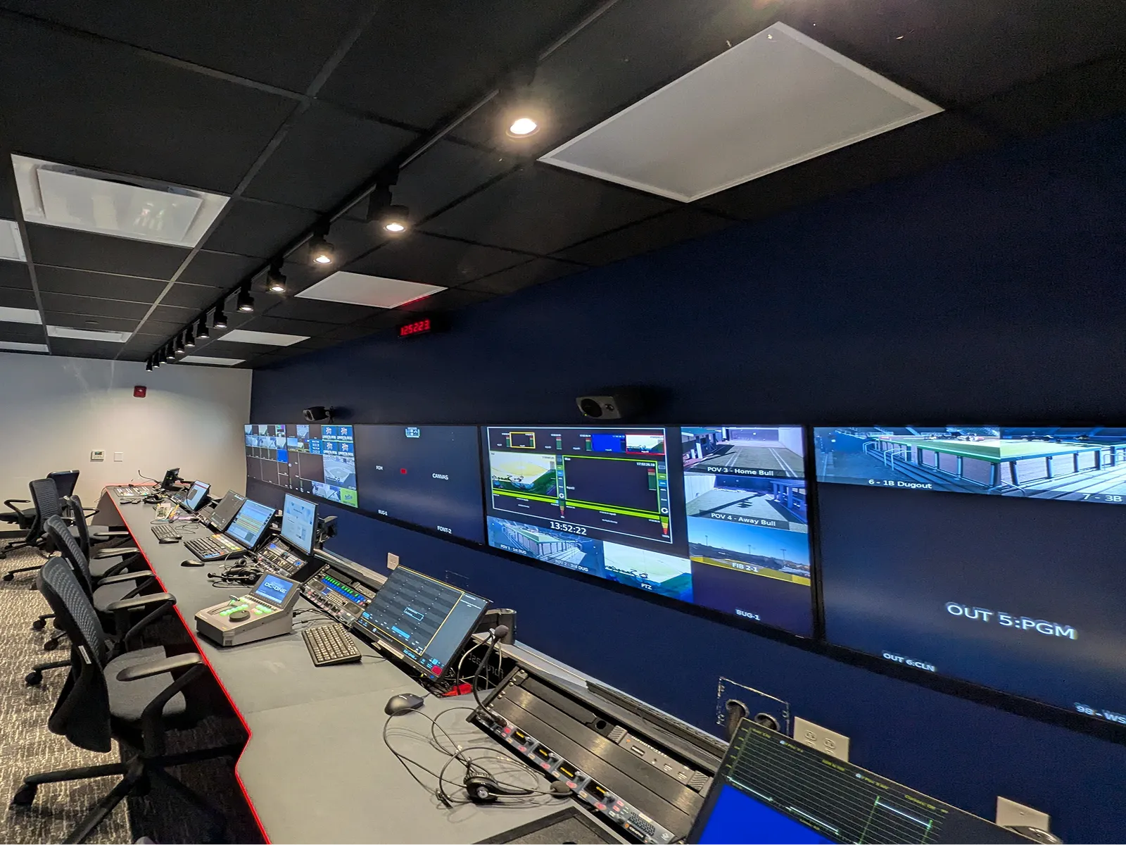 Control room with multiple monitors displaying security and surveillance footage along a long desk with chairs and computer equipment.