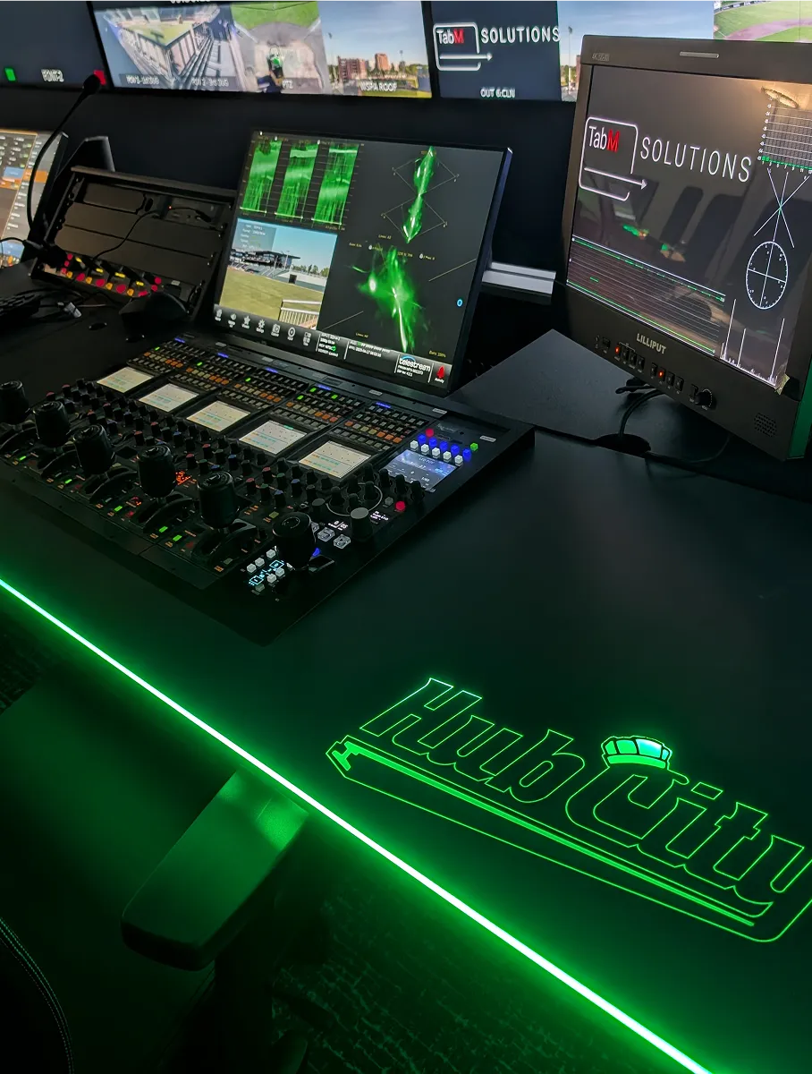 Broadcast control desk with multiple screens displaying surveillance and technical interfaces, featuring green lighting and 'Hub City' logo on the desk.