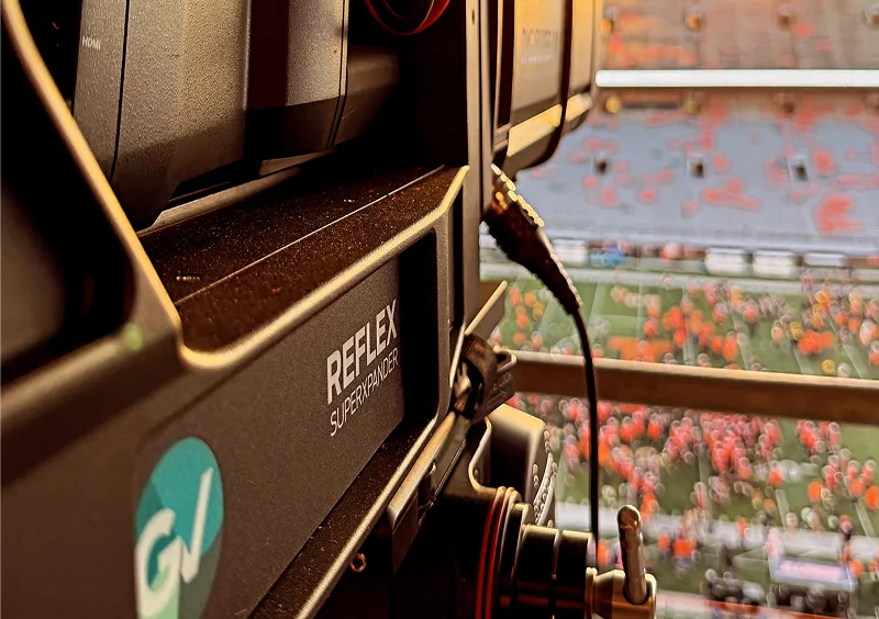 Close-up of a REFLEX SUPERHANDLER camera inside a stadium overlooking a blurred football field and crowd.